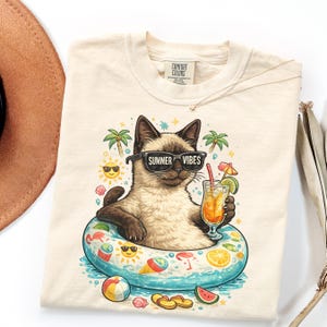 May include: Cream-colored t-shirt featuring a cartoon cat wearing sunglasses with the text "Summer Vibes." The cat is in a pool float with a drink. Palm trees, suns, and other summer-themed graphics surround the cat.