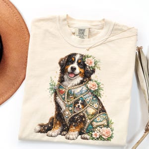 May include: Cream-colored Comfort Colors t-shirt featuring a detailed illustration of a Bernese Mountain Dog. The dog is adorned with floral accents and a decorative garment. A delicate necklace rests on the shirt. The shirt is folded, with a hat and decorative leaves in the background.