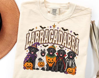 May include: A cream-colored t-shirt featuring a graphic of five black Labrador dogs in Halloween costumes. The text "LABRACADABRA" is printed above the dogs, who are dressed as a wizard, a ghost, a pumpkin, a vampire, and a skeleton with pumpkins.