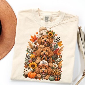 May include: A cream-colored t-shirt with a graphic print of three stacked golden-brown puppies surrounded by autumn elements including sunflowers, pumpkins, pinecones, and fall leaves. The top puppy wears a beanie, the middle a scarf, and the bottom a cap.