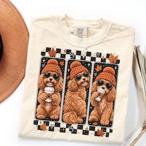 May include: A cream-colored t-shirt featuring a graphic print of three brown curly-haired dogs wearing orange knit beanies and heart-shaped sunglasses. Each dog holds a white coffee cup with a paw print. The design includes autumn leaves.