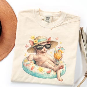 May include: Cream-colored t-shirt featuring a cartoon cat wearing sunglasses and a straw hat, lounging on a pool float with a drink. The text "SUMMER VIBES" is on the sunglasses. The float has a colorful design with suns, watermelon slices, and other summer elements.