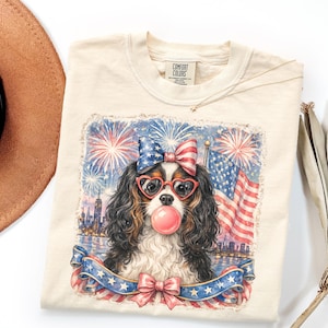 May include: Cream-colored t-shirt featuring a patriotic dog illustration. The dog wears heart-shaped glasses and a bow, blowing a bubblegum bubble. The design includes fireworks, an American flag, and a banner with stars.