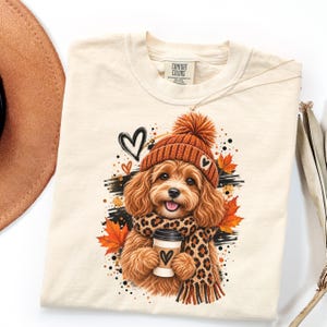 May include: A cream-colored t-shirt featuring a graphic of a fluffy brown dog wearing an orange knit beanie and a leopard print scarf. The dog holds a coffee cup with a heart icon. The design includes orange autumn leaves and black heart outlines.