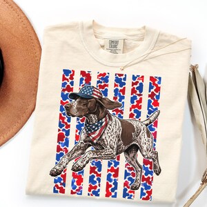 May include: Cream-colored t-shirt featuring a running dog wearing a patriotic hat and bandana. The dog is brown and white, set against a red, white, and blue striped background. The shirt has a Comfort Colors tag.