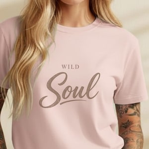 May include: A pale pink t-shirt featuring the text "WILD Soul" in a cursive script. The top has a round neck and is a plain colour. The model is wearing blue jeans.