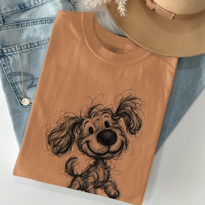 Può includere: Una t-shirt a girocollo color arancione chiaro tinta unita con un'illustrazione a linee nere di un cucciolo arruffato e sorridente con le orecchie cadenti. La maglia è sovrapposta a jeans azzurri, vicino a un cappello beige e una collana.