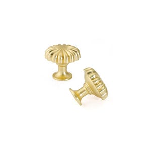 May include: Two gold-colored cabinet knobs with a fluted, flower-like design. The knobs have a rounded top and a short, cylindrical base. One knob is upright, while the other is tilted on its side, both against a white background.