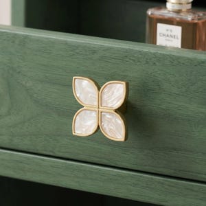 May include: A decorative cabinet knob with a four-petal design. The knob features a gold-colored metal frame with a white, marbled, shell-like inlay. The knob is mounted on a green wooden surface.