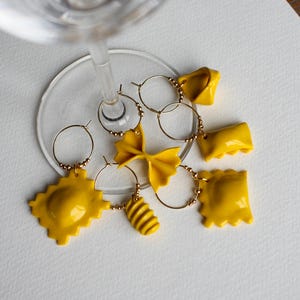Puede incluir: Un conjunto de colgantes para vino con forma de pasta amarilla, incluyendo ravioles, farfalle y rotini, colgados de aros dorados. Los colgantes están dispuestos alrededor de la base de una copa de vino, creando una estética lúdica y única.