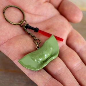 Op de afbeelding: Een groene, knoedelvormige sleutelhanger met een rood lichtzwaard en een zwarte handgreep. De sleutelhanger is bevestigd aan een bronzen sleutelring. De knoedel heeft een glanzende afwerking en is lichtgroen.