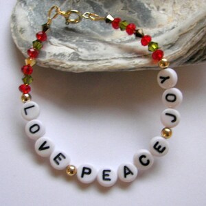 May include: A white beaded bracelet with gold beads and red and green crystal beads. The beads spell out the words "LOVE PEACE JOY".