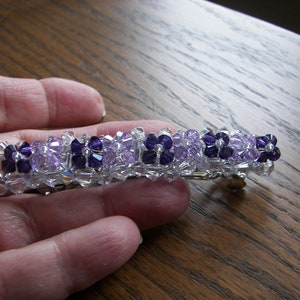 May include: A rectangular hair barrette featuring a repeating pattern of dark purple and light lavender crystal beads arranged in flower-like clusters, interspersed with clear, faceted crystal beads along the entire length of the accessory.