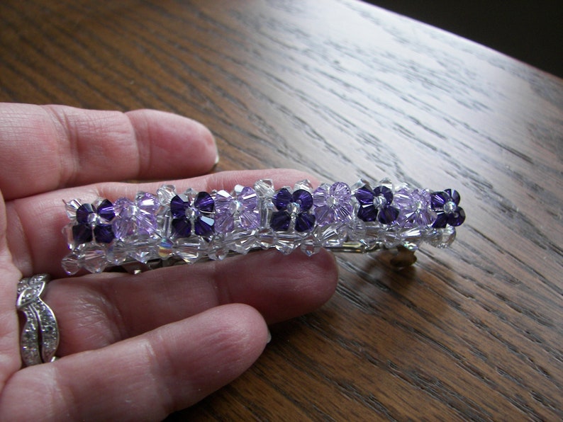 May include: A rectangular hair barrette featuring a repeating pattern of dark purple and light lavender crystal beads arranged in flower-like clusters, interspersed with clear, faceted crystal beads along the entire length of the accessory.