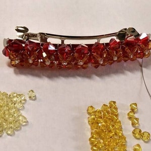 May include: A silver-toned metal hair barrette adorned with a row of faceted red and orange crystal beads arranged in a floral pattern. Below the barrette are two separate piles of loose, faceted yellow and light yellow crystal beads.