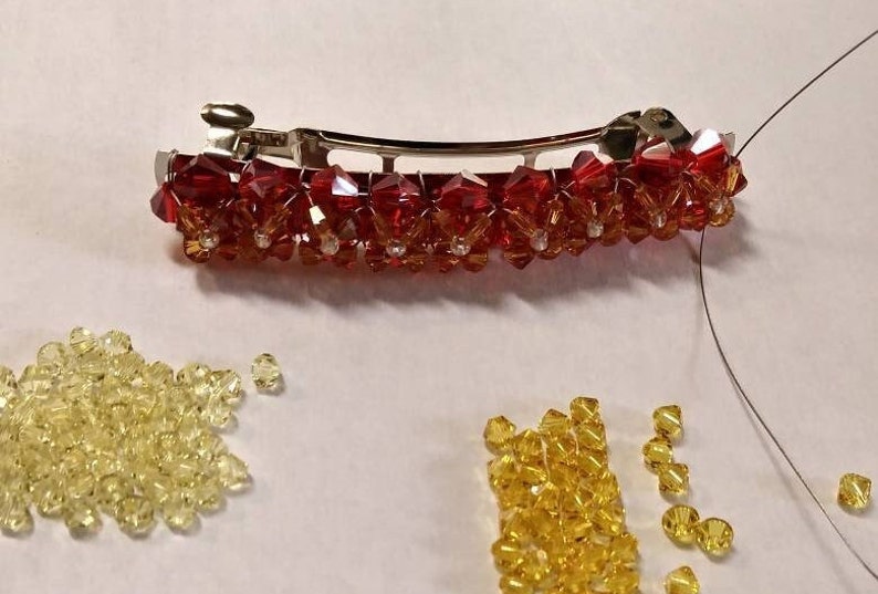 May include: A silver-toned metal hair barrette adorned with a row of faceted red and orange crystal beads arranged in a floral pattern. Below the barrette are two separate piles of loose, faceted yellow and light yellow crystal beads.