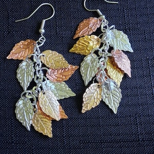 May include: A pair of dangle earrings featuring multiple metallic leaf-shaped charms in copper, gold, and silver tones. Each earring is suspended from a silver-toned hook and consists of several textured leaves attached to small circular metal rings.