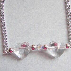 May include: A silver chain necklace with two clear crystal heart charms and a smaller crystal charm in the center.