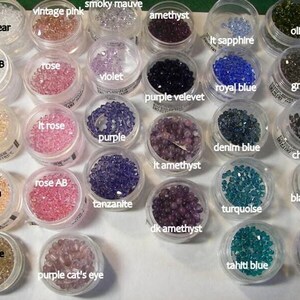 May include: A collection of small, round, faceted beads organized in clear plastic containers. Each container is labeled with color names including clear, vintage pink, smoky mauve, amethyst, lt sapphire, olive, clear AB, rose, violet, purple velevet, royal blue, greige, silk, lt rose, purple, denim blue, chrome, peach, rose AB, tanzanite, lt amethyst, turquoise, black diamond, champagne, purple cat's eye, dk amethyst, tahiti blue, and jet black.