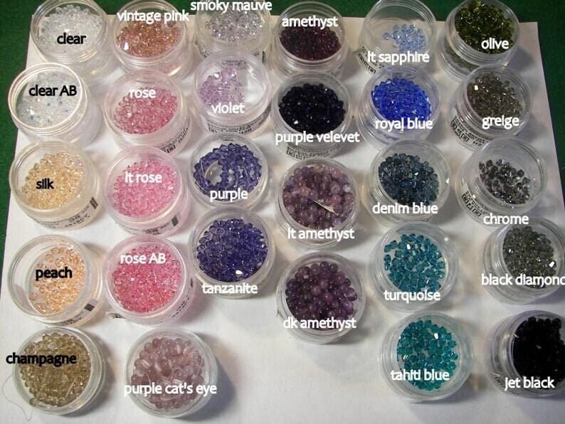May include: A collection of small, round, faceted beads organized in clear plastic containers. Each container is labeled with color names including clear, vintage pink, smoky mauve, amethyst, lt sapphire, olive, clear AB, rose, violet, purple velevet, royal blue, greige, silk, lt rose, purple, denim blue, chrome, peach, rose AB, tanzanite, lt amethyst, turquoise, black diamond, champagne, purple cat's eye, dk amethyst, tahiti blue, and jet black.