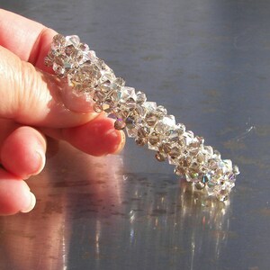 May include: A cylindrical hair accessory crafted from numerous small, clear, faceted crystals arranged in a dense, textured pattern. The crystals reflect light, creating a shimmering effect across the entire surface of the tubular hair clip.