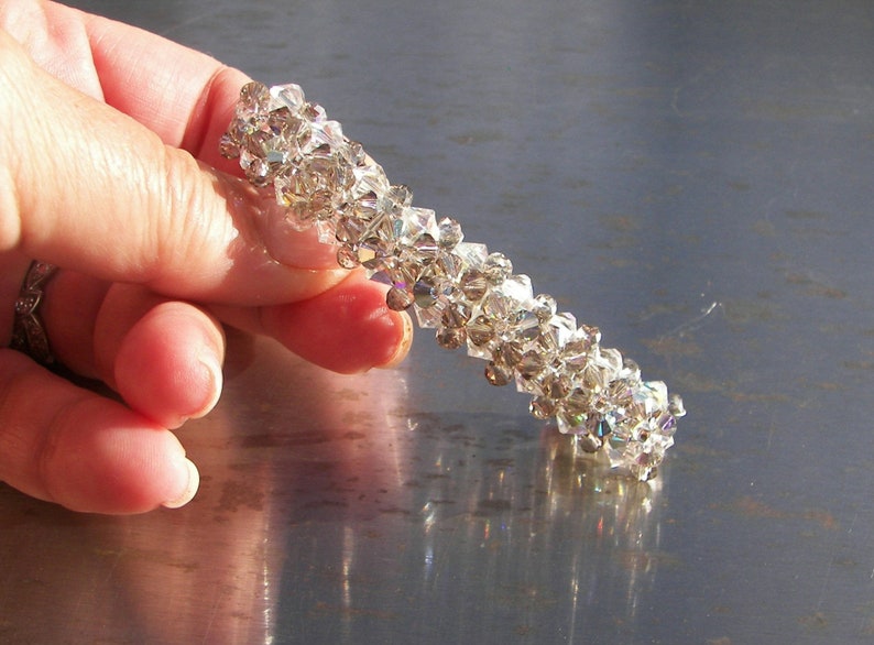 May include: A cylindrical hair accessory crafted from numerous small, clear, faceted crystals arranged in a dense, textured pattern. The crystals reflect light, creating a shimmering effect across the entire surface of the tubular hair clip.