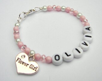 May include: A pink and white beaded bracelet with a silver heart charm that says "Flower Girl". The bracelet has white beads with the name "Olivia" spelled out in black letters.