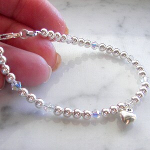 May include: Silver beaded bracelet with a small heart charm and clear crystal accents. The bracelet is held up against a white marble surface. The bracelet is approximately 7 inches in length.