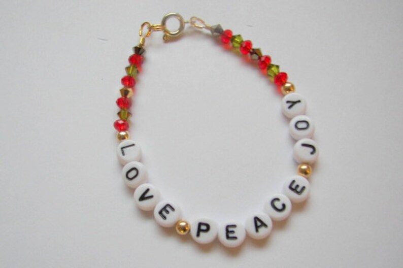 May include: A white beaded bracelet with gold spacers and red and green crystal beads. The beads spell out the words "LOVE PEACE JOY".