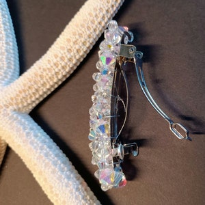 May include: A hair clip featuring a cluster of clear, iridescent, multifaceted crystal beads attached to a silver-toned metal barrette mechanism. The beads reflect light with rainbow hues, and the metal clip has a hinged, spring-loaded closure.