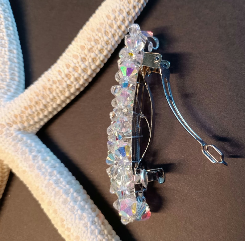 May include: A hair clip featuring a cluster of clear, iridescent, multifaceted crystal beads attached to a silver-toned metal barrette mechanism. The beads reflect light with rainbow hues, and the metal clip has a hinged, spring-loaded closure.