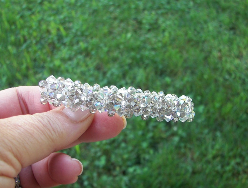 May include: A hair barrette featuring a dense arrangement of multifaceted, clear, iridescent crystal beads. The beads are tightly clustered together to form a cylindrical shape, reflecting light with a spectrum of colors across the surface.