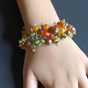 May include: A cluster bracelet featuring a variety of round beads in shades of orange, olive green, and metallic bronze. The beads are attached to a silver-toned metal chain with small clear glass spacer beads interspersed throughout the design.