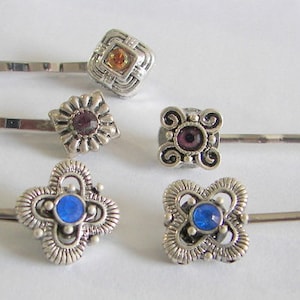 May include: Set of six silver hair pins with decorative tops. Each pin features a different design with a jeweled center. The jewels are purple, orange, and blue.