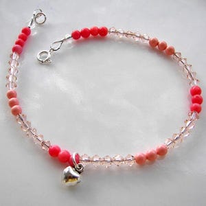 May include: A delicate bracelet featuring alternating sections of bright coral-colored round beads, soft peach-colored round beads, and clear faceted crystal beads. The piece includes a small silver-toned heart charm and a silver-toned spring ring clasp.