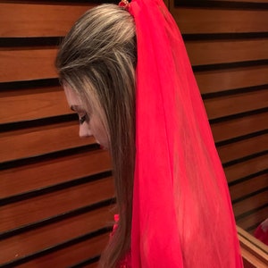 May include: A long, vibrant red sheer veil attached to a hair accessory. The veil is made of a lightweight, translucent fabric that drapes down from the crown of the head, creating a flowing effect. The accessory securing the veil is visible.