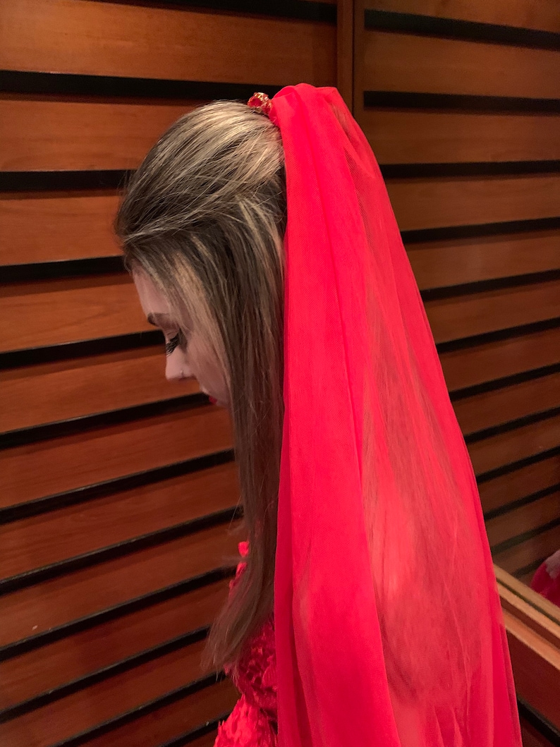 May include: A long, vibrant red sheer veil attached to a hair accessory. The veil is made of a lightweight, translucent fabric that drapes down from the crown of the head, creating a flowing effect. The accessory securing the veil is visible.