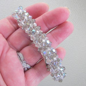 May include: A rectangular hair barrette featuring a dense arrangement of multifaceted, clear, and iridescent crystal beads. The beads are tightly clustered together to create a textured, reflective surface that catches light, forming a long, narrow accessory.