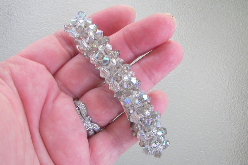 May include: A rectangular hair barrette featuring a dense arrangement of multifaceted, clear, and iridescent crystal beads. The beads are tightly clustered together to create a textured, reflective surface that catches light, forming a long, narrow accessory.