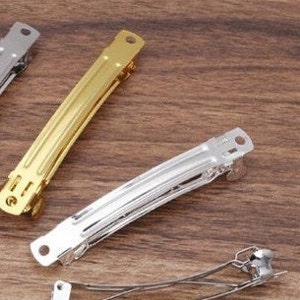 May include: A collection of three metal hair barrettes featuring a hinged clasp mechanism. One barrette is finished in a polished gold tone, while the other two are finished in a bright silver tone. Each barrette has a long, slender rectangular body.