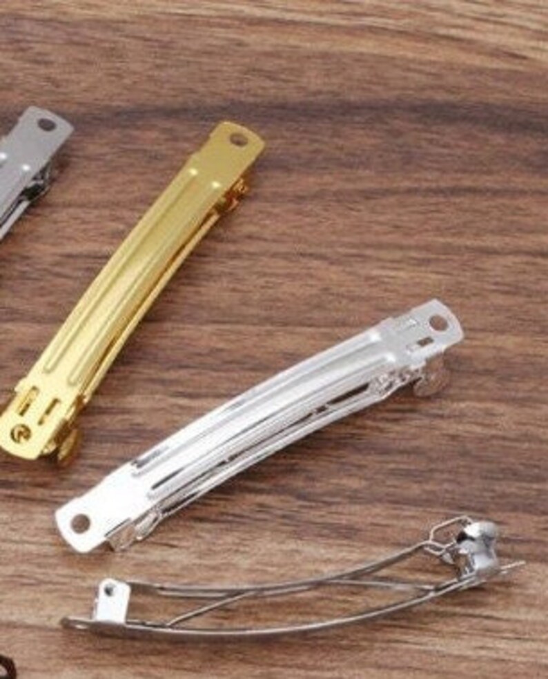 May include: A collection of three metal hair barrettes featuring a hinged clasp mechanism. One barrette is finished in a polished gold tone, while the other two are finished in a bright silver tone. Each barrette has a long, slender rectangular body.