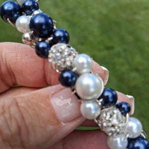 May include: A beaded bracelet featuring navy blue, white, and silver-colored beads. The bracelet has a mix of pearl-like and rhinestone-encrusted beads, creating a textured and elegant design. The bracelet is held against a blurred green background.