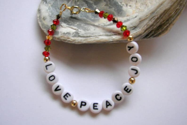 May include: A white beaded bracelet with gold beads and red and green crystal beads. The beads spell out the words "LOVE PEACE JOY".