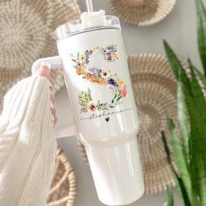 May include: White tumbler featuring a floral "S" design and the name "Stephanie" in script. The tumbler has a handle and a clear lid with a straw. The floral design includes orange, purple, and yellow flowers. A small black heart is below the name.