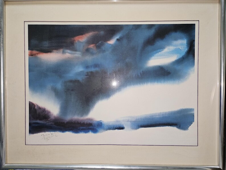 May include: Abstract watercolor painting in a silver frame. The artwork features a dramatic sky with shades of blue, black, and hints of pink. The bottom of the painting has a dark blue and purple landscape.