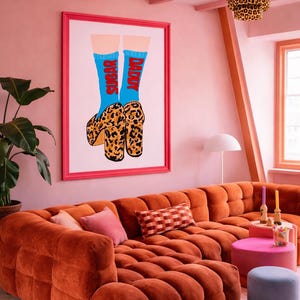 Op de afbeelding: Een ingelijst kunstwerk met benen in blauwe sokken met de woorden "SUGAR" en "DADDY" in het rood, gecombineerd met plateauzolen met luipaardprint. Het kunstwerk hangt aan een roze muur boven een oranje hoekbank.