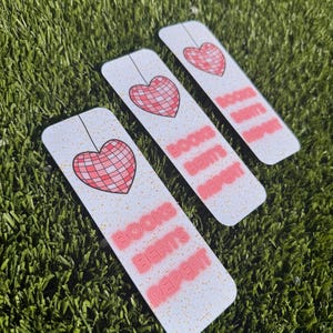 May include: Three white bookmarks with a pink heart design and the text "BOOKS BEATS REPEAT" in a retro font. The bookmarks are arranged on a bed of green artificial grass, creating a visually appealing display.
