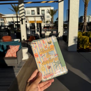 Può includere: Una custodia trasparente per telefono con un design colorato con il testo "Booked & Sunburn". La custodia ha uno sfondo a righe blu chiaro e giallo con illustrazioni tropicali, tra cui un furgone rosso, fiori e noci di cocco.