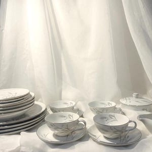 May include: A white porcelain tea set with a delicate silver leaf design. The set includes cups, saucers, plates, a sugar bowl, and a creamer. The edges of the pieces are trimmed with silver. The set is arranged on a white surface.