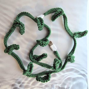 Könnte beinhalten: Ein grünes, gehäkeltes USB-Kabel, das wie eine Weinrebe mit Blättern geformt ist. Das Kabel hat weiße USB-Anschlüsse an beiden Enden. Das handgefertigte Kabel ist eine einzigartige und dekorative Möglichkeit, Geräte aufzuladen.